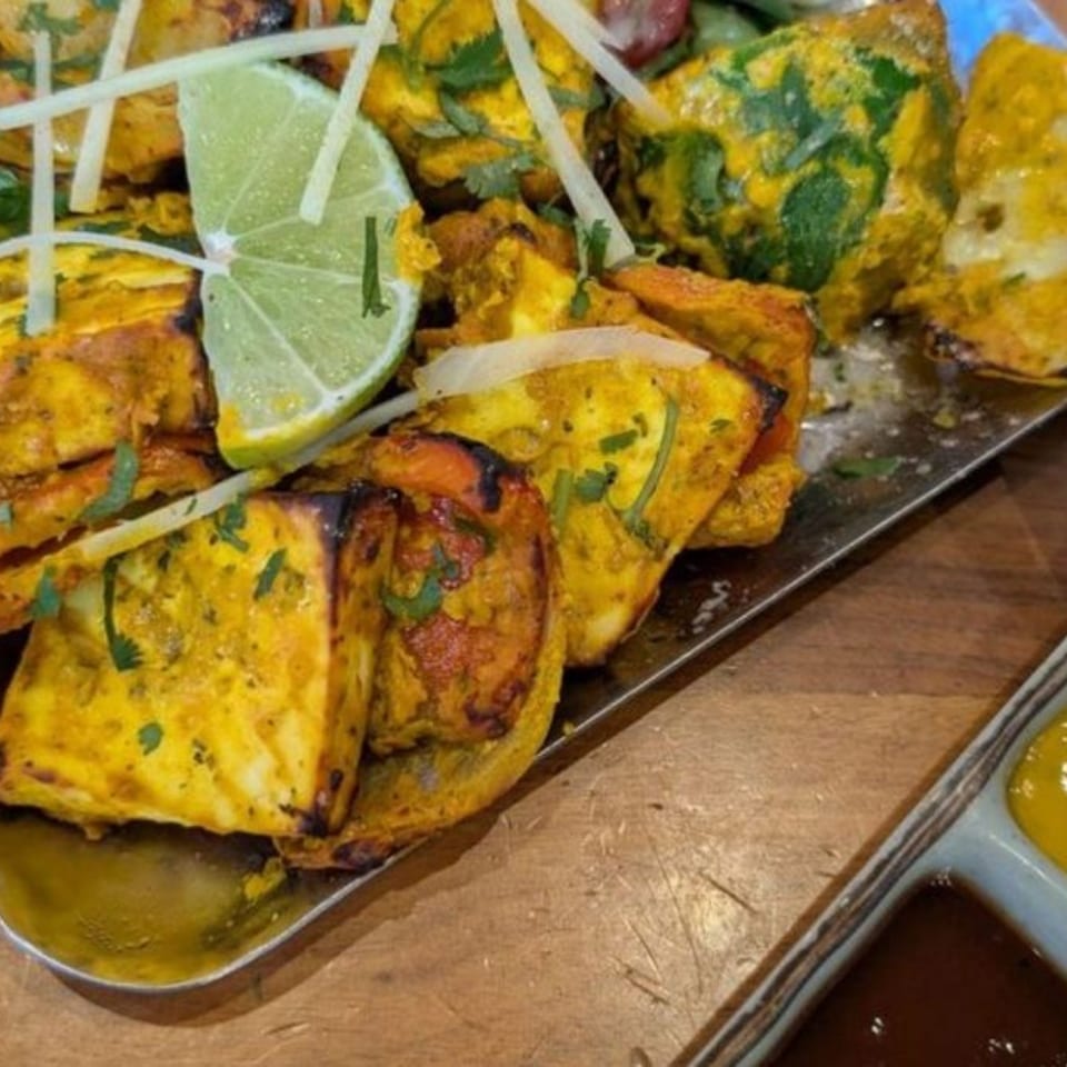 Golden grilled paneer and vegetables, garnished with lime and cilantro, served on a metal platter with sauces.